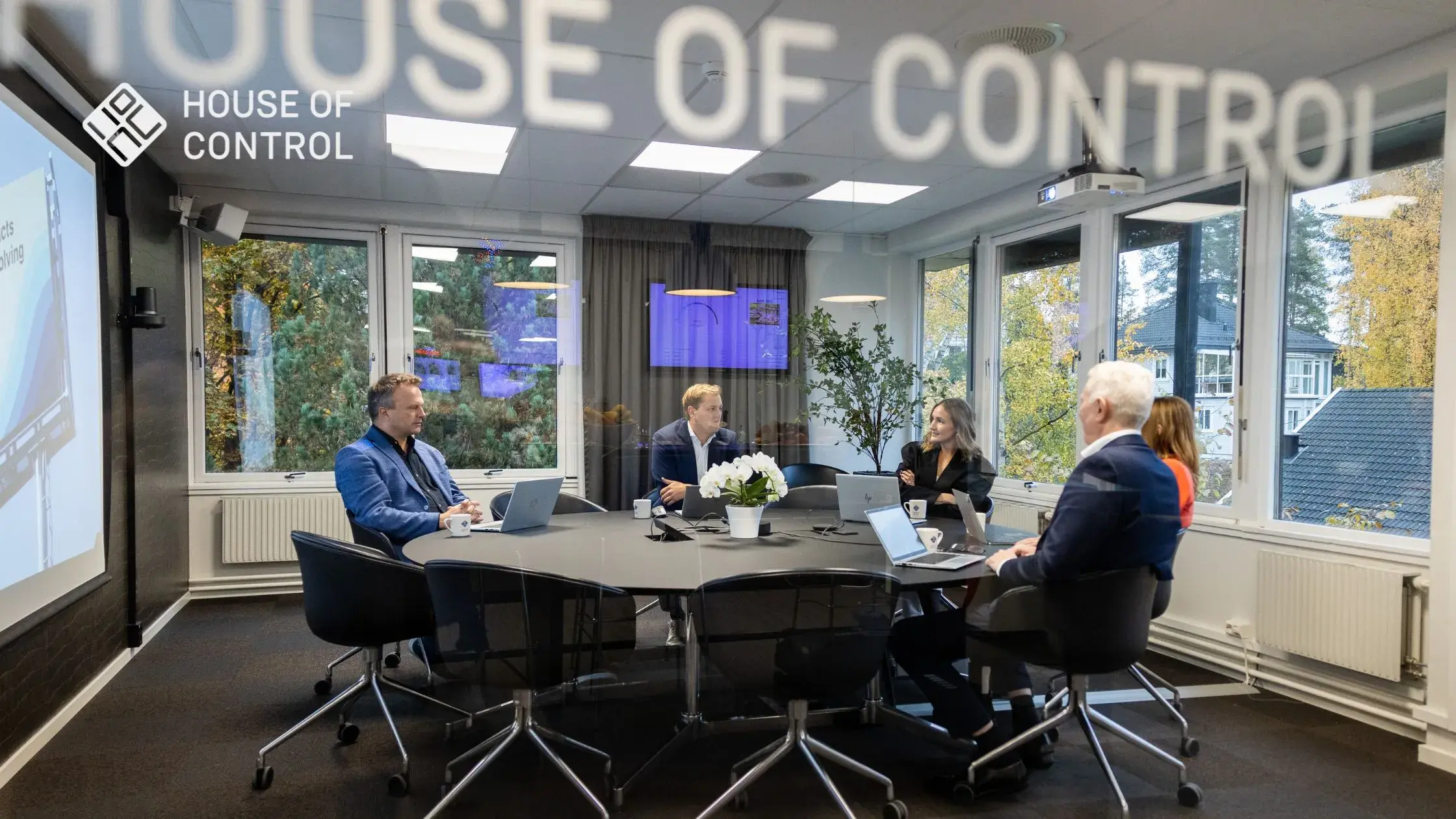 A group of employees sitting down in a meeting, in a big room, at a round table. The House of Control logo is at the right side top